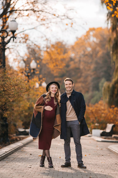 Young stylish couple in love at an autumn city park. Happy stylish man and beautiful pregnant woman in modish dress and hat posing outdoors on an autumn day - 写真・画像
