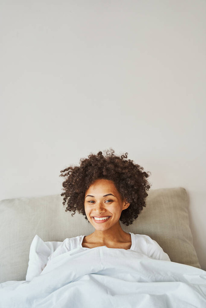 Curly-headed young woman with a radiant smile resting in bed - Foto, Imagem