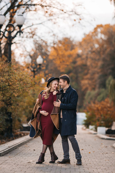 A couple of beautiful lovers spend time outdoors on the fall day. Pretty woman in a red dress and modish hat and a handsome man in a raincoat posing at the city park on a sunny autumn day - Foto, afbeelding