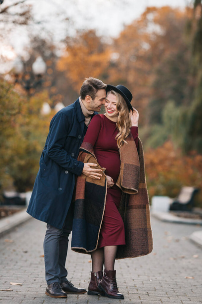 Happy young parents, a stylish man in a raincoat and a pregnant young woman in red dress and fashionable hat walking together in an autumn city park on an autumn day - Fotografie, Obrázek