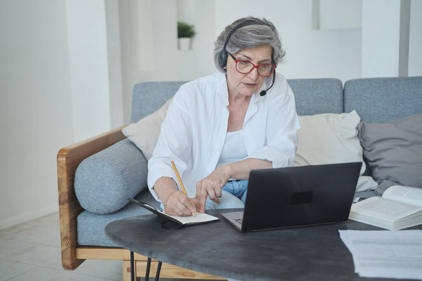Smart and motivated retired old woman using laptop and earphones to learning new skills online - 写真・画像