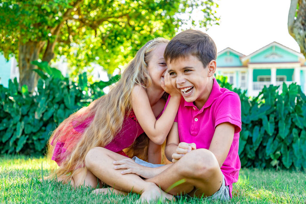 Маленька дівчинка шепоче анекдот у вухо хлопчика, хлопчик сміється, літній фон саду
 - Фото, зображення