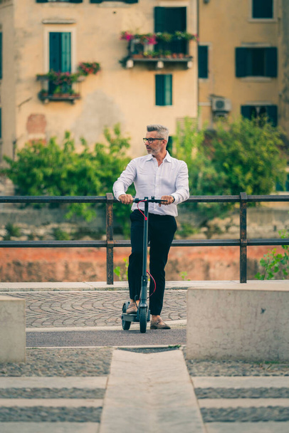 Şehirde işe giderken elektrikli scooter kullanan takım elbiseli yakışıklı bir iş adamı. Ekolojik ulaşım kavramı - Fotoğraf, Görsel