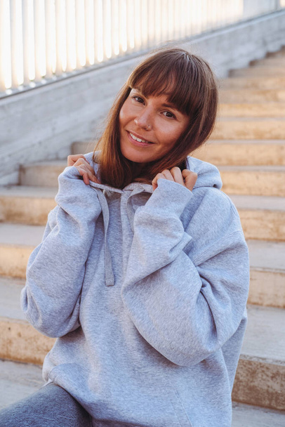 Beautiful woman in grey sweatshirt seats on a stairs. - Фото, зображення