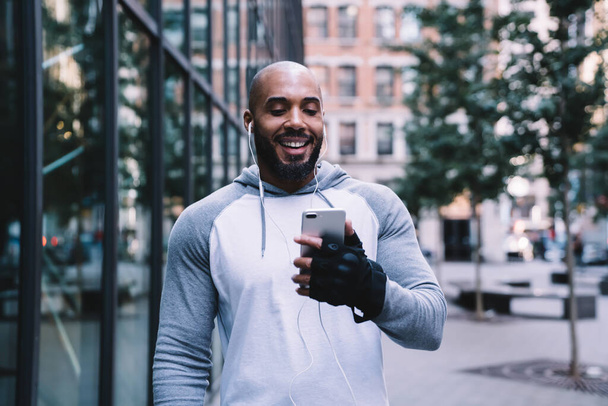 Athletic bald bearded African American guy in sportswear using smartphone and smiling while standing on street near modern office building and listening to music - Zdjęcie, obraz