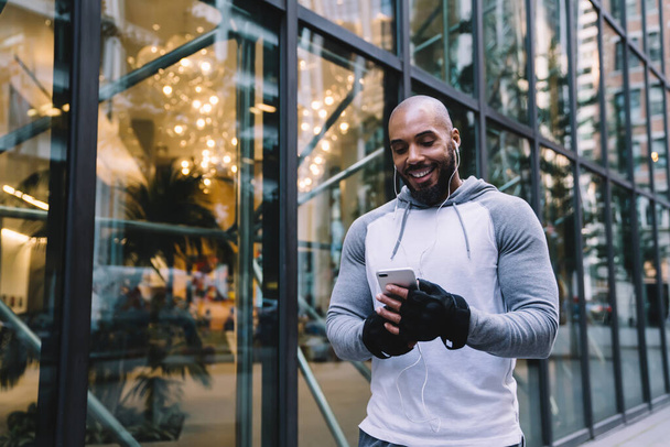 Cheerful bald bearded black man in sportswear using smartphone and listening to music during workout on street near modern office building - Фото, зображення