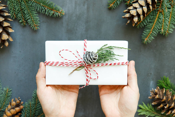 Close up of man hands holding white wrapped gift box with ribbon, sprig of conifer, cone on black background. Winter holiday pattern. Top view. Christmas background. - Fotoğraf, Görsel