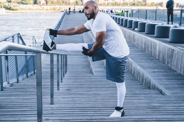 Side view of athletic African American man practicing on waterfront stretching legs on metal fence looking away against urban background  - Zdjęcie, obraz