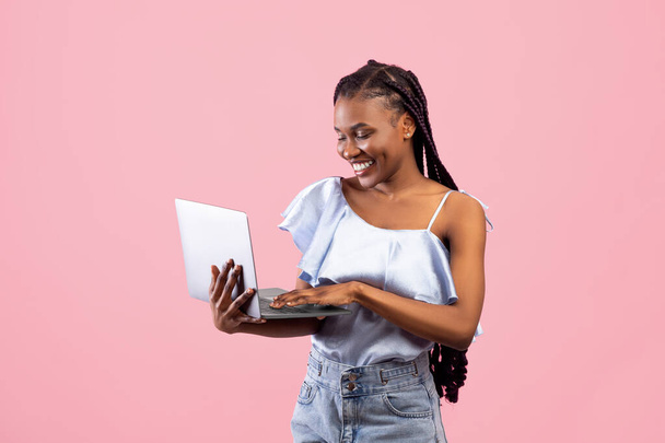 Young black woman with afro bunches using laptop, freelancing, browsing web on pink studio background - Foto, Imagem