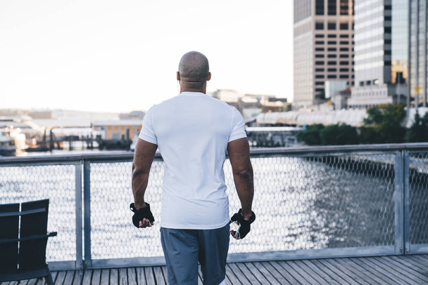Back view of anonymous bald black male in sportswear looking at river and downtown while listening to music and standing on embankment - Foto, Imagem