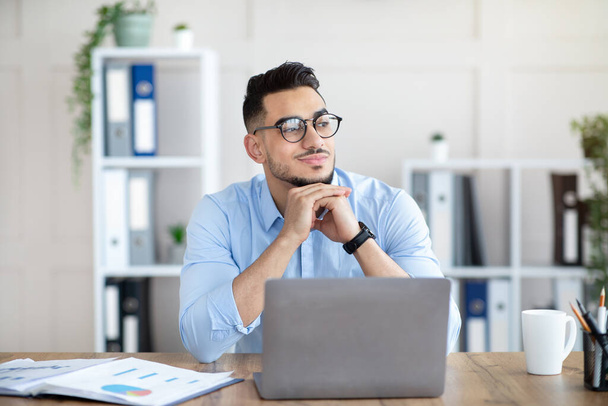 Sonhador árabe cara em óculos sentado no laptop no escritório, olhando para o lado, tendo pausa do trabalho on-line - Foto, Imagem