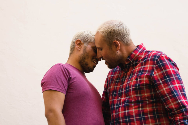 Hombres adultos con ropa casual abrazándose y besándose mientras están de pie contra el fondo beige durante la fecha - Foto, imagen