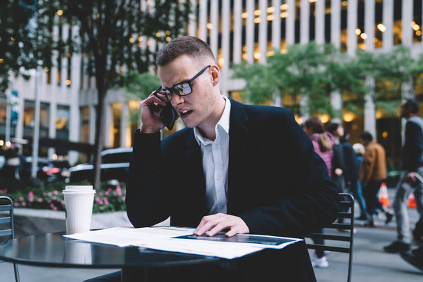 Angry entrepreneur wearing black office suit and glasses sitting at table in street cafe talking on phone looking over paperwork  - Foto, imagen