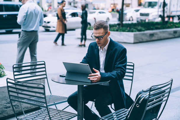 Handsome concentrated man in business suit and glasses browsing and watching tablet attentively while sitting at table in street cafe - Photo, Image