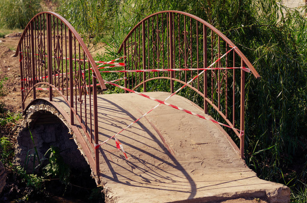 Eine kleine Brücke über den Fluss mit Absperrband. Rotes und weißes Band verbietet das Überqueren der Brücke. Sommernachmittage. Selektiver Fokus. - Foto, Bild