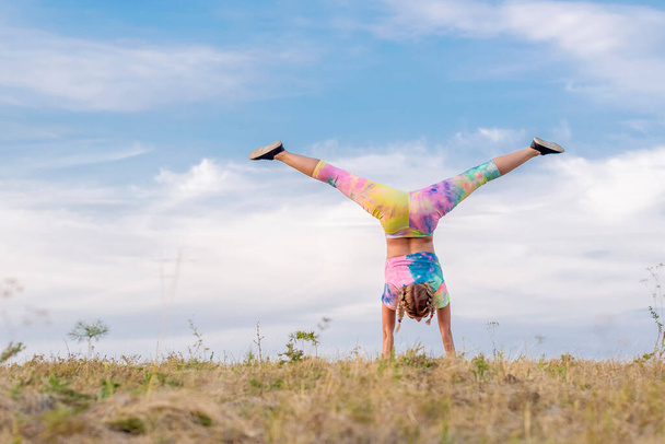 Young girl in a bright tracksuit performs a handstand against the blue summer sky. Legs spread apart - split. Outdoor gymnastics. - Фото, изображение