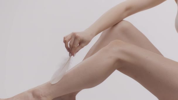 Moderna depilación. Primer plano de señora caucásica irreconocible acariciando la piel lisa de sus piernas perfectas con pluma - Imágenes, Vídeo