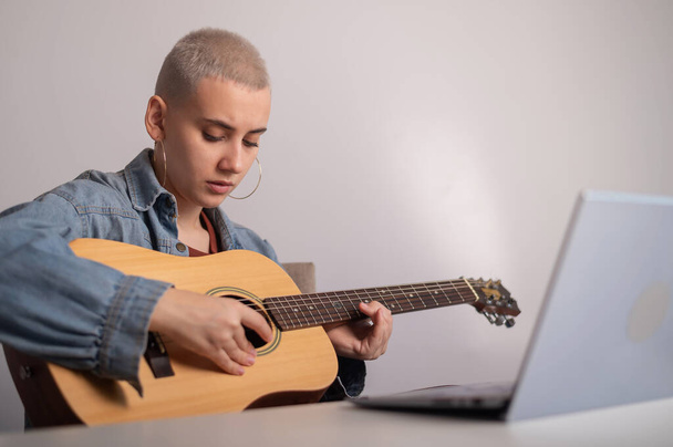 Young caucasian woman with short blonde hair playing guitar and watching training video on laptop. - Фото, изображение