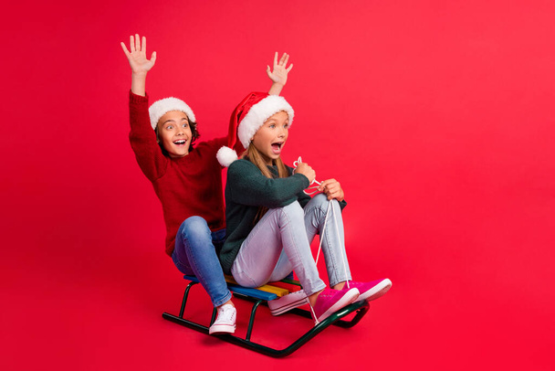 Photo of sweet excited schoolkids dressed sweaters riding sledges rising hands looking empty space isolated red color background - 写真・画像