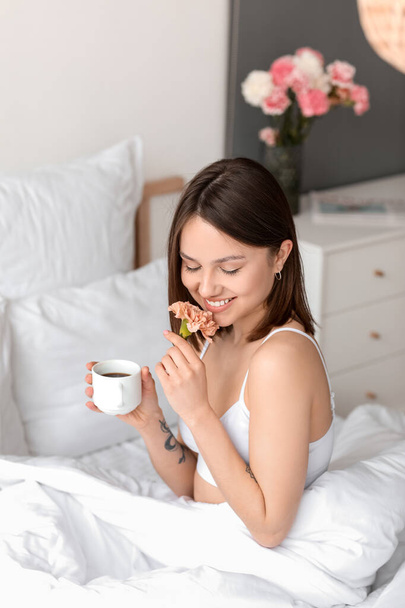 Young woman with cup of coffee and flower in bedroom - Foto, afbeelding