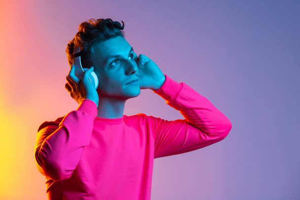 Retrato de mitad de longitud del joven hombre caucásico en auriculares aislados color púrpura fondo de estudio en filtro de luz de neón rosa. Concepto de emoción humana, expresiones faciales, juventud, sentimientos, ad. - Foto, imagen