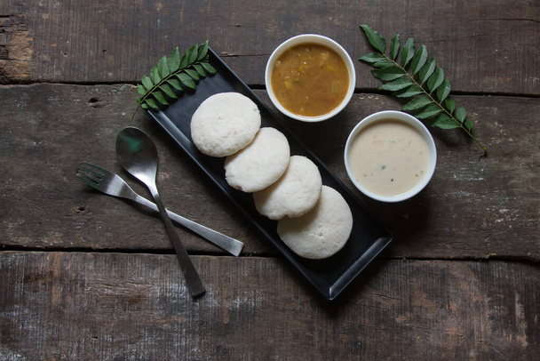 South Indian snacks idli sambar or idly sambhar prepared by steaming fermented rice and served with coconut dip and vegetable soup in a black tray. Top view. - Photo, Image
