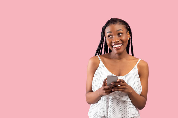 Álmodozó fekete nő használ okostelefon, nézi félre szabad helyet rózsaszín stúdió háttér - Fotó, kép