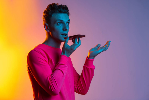 Half length portrait of young Caucasian man with phone, gadget isolated purple color studio background in pink neon light filter. Concept of human emotion, facial expressions, youth, feelings, ad. - Φωτογραφία, εικόνα
