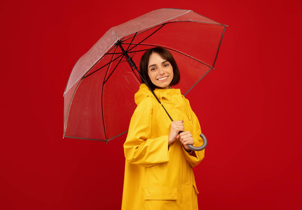 Koncept podzimní nálady. Šťastná dáma stojí pod průhledným deštníkem, usmívá se a dívá se na kameru přes červené pozadí - Fotografie, Obrázek
