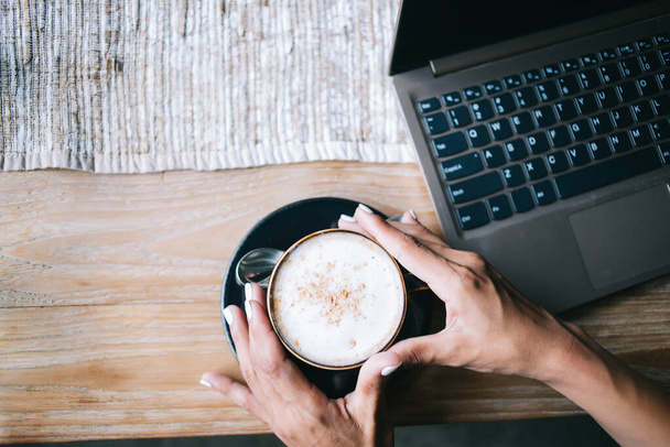 Top view of anonymous dark-skinned woman with black cup of cappuccino on saucer with teaspoon on wooden table with gray cloth and laptop - Foto, afbeelding