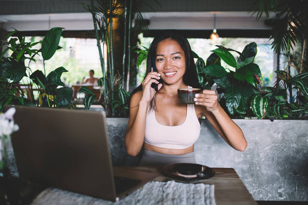 Cheerful beautiful Asian woman sitting at table at restaurant talking on phone holding cup of coffee in hand looking at camera - Fotografie, Obrázek