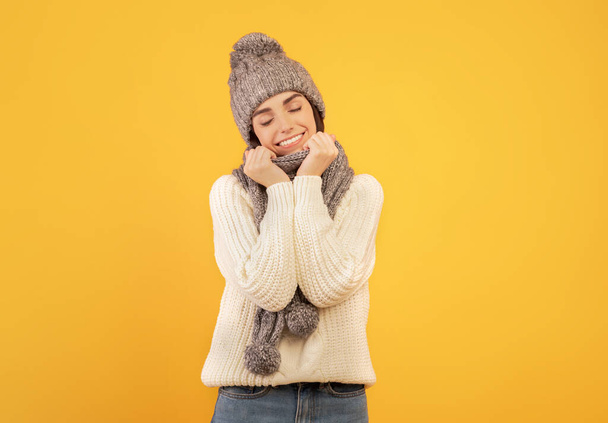 Happy pretty lady wearing white knitted sweater, grey scarf and hat, standing with closed eyes over yellow studio background - Fotografie, Obrázek