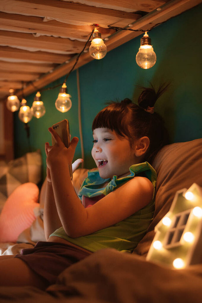 Little happy girl with funny hairstyle sitting in childrens room on her bunk bed holding smartphone - Photo, Image