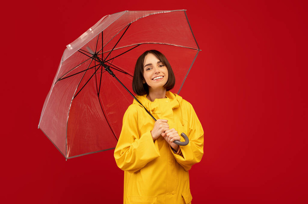 Millennial donna caucasica in posa con ombrello trasparente, sorridente e guardando la fotocamera su sfondo rosso - Foto, immagini