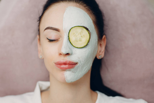 Young woman applying mask of clay on face with cucumber on eyes in beauty spa - Foto, Imagem