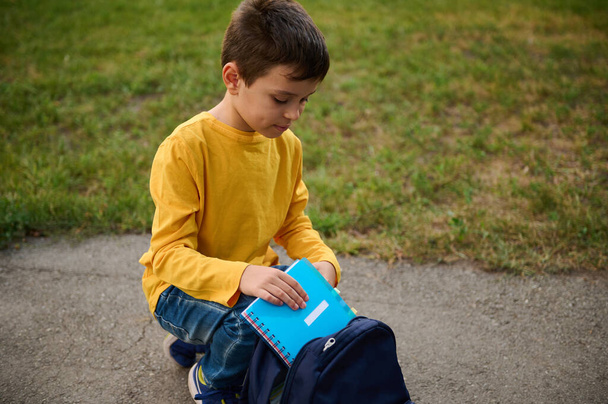 Очаровательный красивый школьник в начальной школе сидит на коленях на дорожке в парке, кладет блокнот и пенал в рюкзак, возвращаясь домой после стрельбы. Снова в школу - Фото, изображение