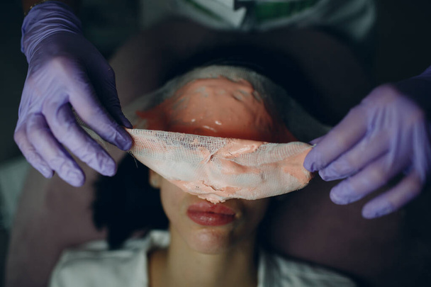 Alginate processus de suppression de masque facial. Les mains d'une esthéticienne tenant une spatule. Femme allongée sur un canapé dans un bureau de cosmétologue. Soins du visage dans le salon de beauté spa - Photo, image