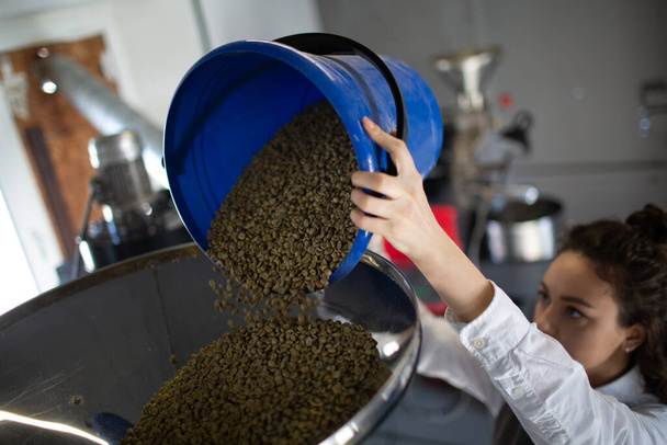 Coffee roaster machine at coffee roasting process. Young woman worker barista loading green coffee beans in apparatus - Photo, Image