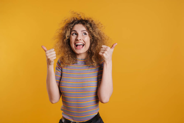 Happy excited young casual woman with fizzy hairstyle standing over yellow wall background, giving thumbs up - Foto, afbeelding