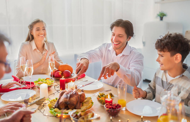 Wesoły młodzieniec świętujący Boże Narodzenie lub Święto Dziękczynienia w domu, krojący pieczonego indyka na uroczystą rodzinną kolację - Zdjęcie, obraz