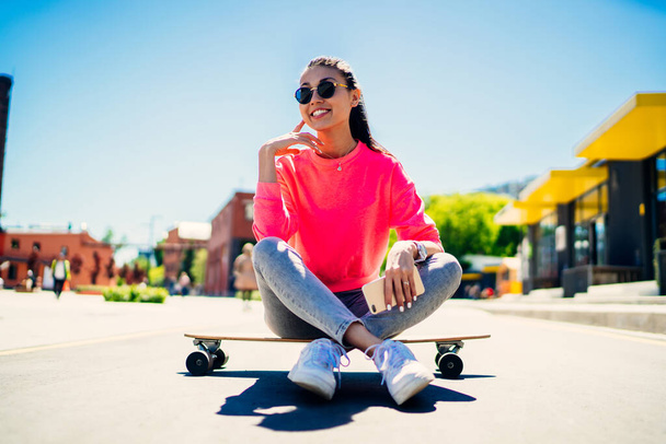 Cute Asian female in trendy sunglasses and streetwear resting at longboard and dreaming about skater career, happy hipster girl in sweatshirt enjoying free time for practicing boarding in city - Фото, изображение