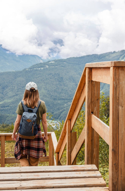 Позаду - молода блондинка з рюкзаком і шовковистою сорочкою, яка дивиться на Кавказькі гори і хмарне небо, що стоять з дерев'яної точки зору, ходячи активним здоровим способом життя, насолоджуючись природою. - Фото, зображення