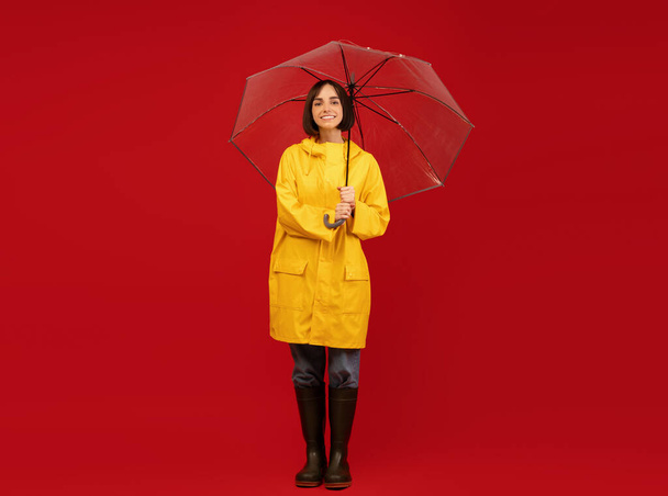Осенний стиль. Студийный снимок женщины тысячелетия в плаще, стоящей под зонтиком и позирующей на красном фоне - Фото, изображение