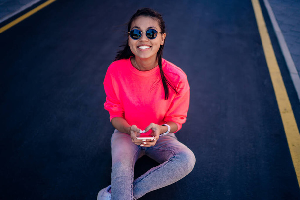Portrait of happy hipster girl in trendy sunglasses and pink sweatshirt with copy space for brand name using modern cellphone device and smiling at camera, cheerful Asian blogger connecting to 4g - Fotografie, Obrázek