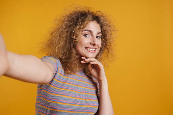 Happy smiling young blonde woman with fizzy hair in casual clothes standing over yellow wall background, taking a selfie - 写真・画像