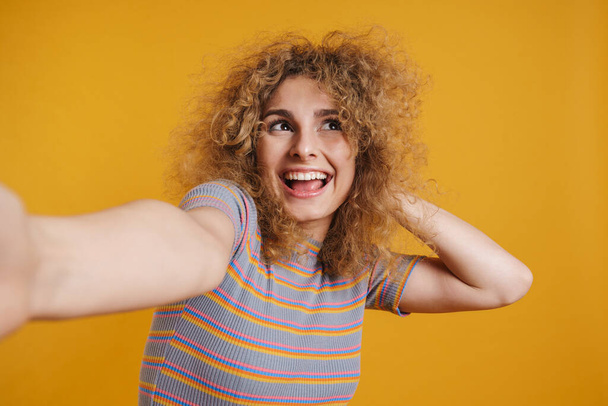 Happy excited young casual woman with fizzy hairstyle standing over yellow wall background, taking a selfie - Valokuva, kuva