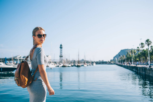 Side view of stylish young woman in casual clothes and sunglasses looking at camera over shoulder while standing near cam water in harbor of Barcelona City - Foto, imagen