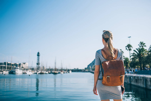 Back view of anonymous female with smartphone and backpack standing near water and admiring harbor with ships on sunny day in Barcelona City - Foto, Imagem