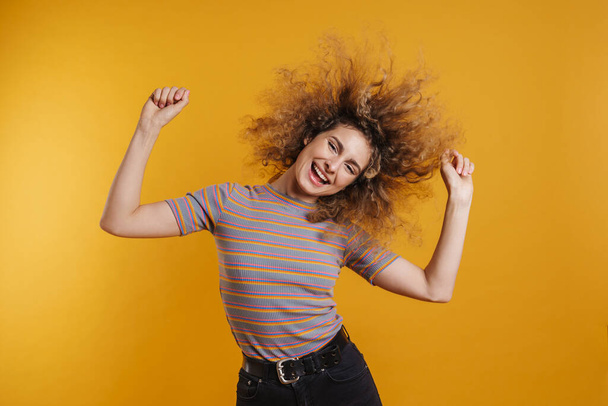 Happy excited young casual woman with fizzy hairstyle standing over yellow wall background, dancing, celebrating - Foto, imagen