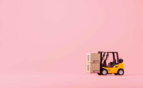 Logistiek, en bezorgservice - Vorkheftruck model en papieren kartons of pakket met een winkelwagen logo op roze achtergrond. Shopping service op het online web en biedt thuisbezorging. met kopieerruimte - Foto, afbeelding